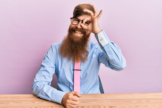 Young irish redhead man wearing business shirt and tie sitting on the table doing ok gesture with hand smiling, eye looking through fingers with happy face.