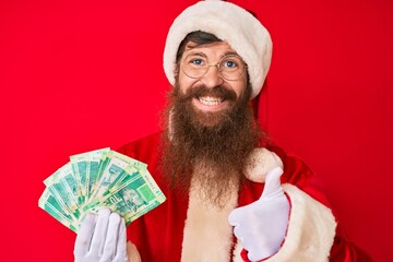 Handsome young red head man with long beard wearing santa claus costume holding african rand smiling happy and positive, thumb up doing excellent and approval sign