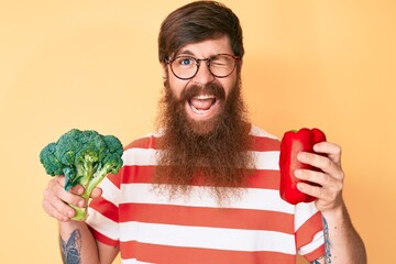 Handsome young red head man with long beard holding broccoli and red pepper winking looking at the...