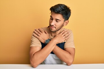 Young handsome man wearing casual clothes sitting on the table hugging oneself happy and positive,...