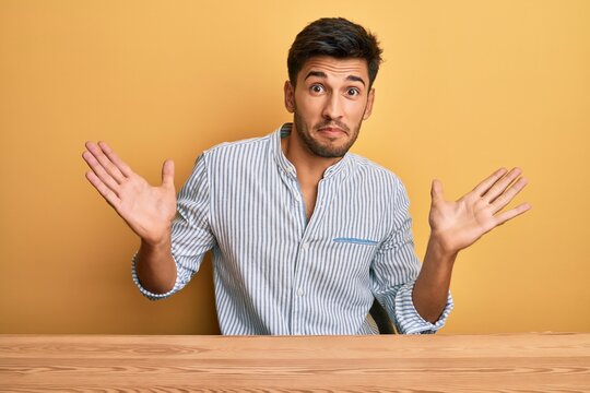 Young handsome man wearing casual clothes sitting on the table clueless and confused with open arms, no idea and doubtful face.