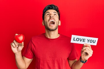 Young handsome man holding red heart and love you message angry and mad screaming frustrated and furious, shouting with anger looking up.