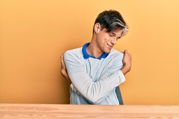 Young hispanic man wearing casual clothes sitting on the table hugging oneself happy and positive, smiling confident. self love and self care