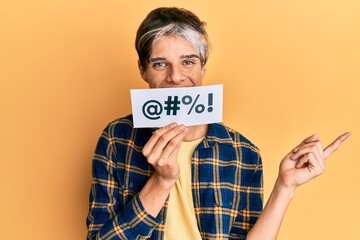 Young hispanic man covering mouth with insult message paper smiling happy pointing with hand and...