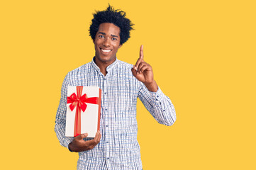 Handsome african american man with afro hair holding gift surprised with an idea or question pointing finger with happy face, number one