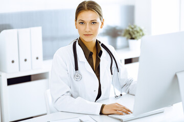 Woman-doctor typing on pc computer while sitting at the desk in hospital office. Physician at work