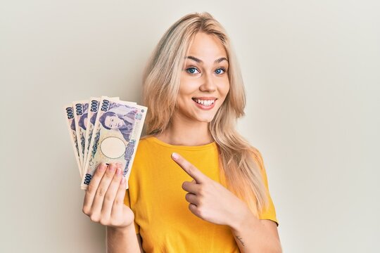 Beautiful caucasian blonde girl holding 5000 japanese yen banknotes smiling happy pointing with hand and finger