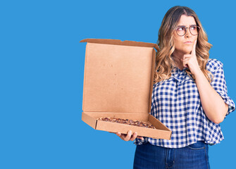 Young caucasian woman holding delivery pizza box serious face thinking about question with hand on chin, thoughtful about confusing idea