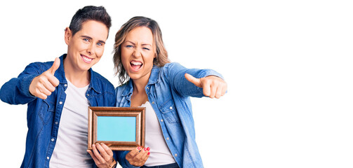 Couple of women holding empty frame approving doing positive gesture with hand, thumbs up smiling and happy for success. winner gesture.