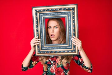 Fototapeta premium Young caucasian woman with blond hair holding empty frame in shock face, looking skeptical and sarcastic, surprised with open mouth