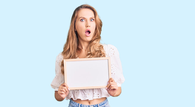 Young caucasian woman with blond hair holding blank empty banner scared and amazed with open mouth for surprise, disbelief face