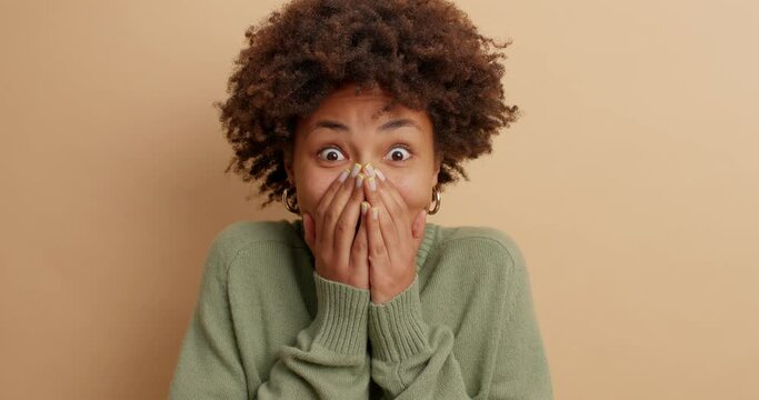 Cute surprised young Afro American woman covers mouth and gazes surprisingly at camera stands speechless cannot believe in unexpected news poses against brown background expresses great wonder