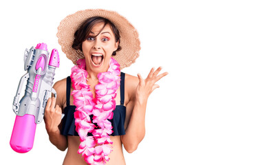 Beautiful young woman with short hair wearing bikini and hawaiian lei holding water gun celebrating victory with happy smile and winner expression with raised hands
