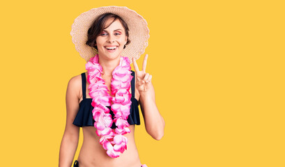 Beautiful young woman with short hair wearing bikini and hawaiian lei smiling with happy face winking at the camera doing victory sign. number two.