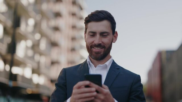 Successful happy caucasian young adult bearded businessman commuting to work using smartphone chatting messages smiling walking the street outdoor. Business people.