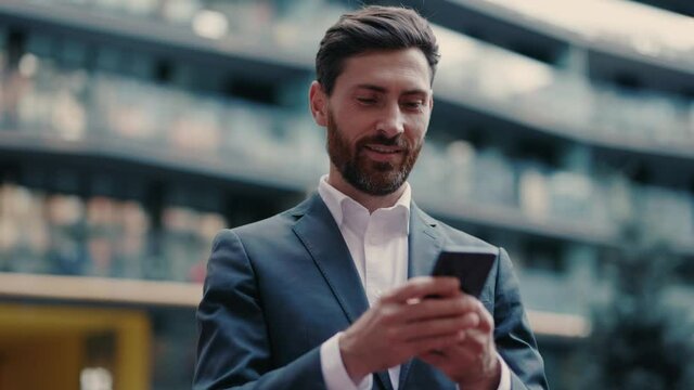Confident positive adult caucasian entrepreneur browsing smartphone app on the street. Handsome and prosperous businessman. Corporate people. Success, luxury, technology.