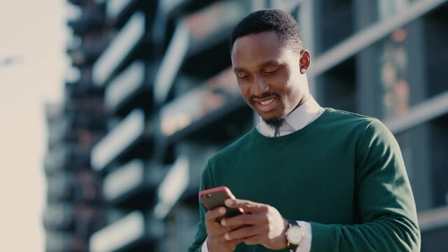 Confident african american man using mobile phone in modern city district. Positive business guy browsing financial news smiling standing outdoor. Successful adult.