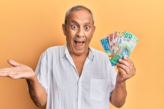 Handsome mature man holding australian dollars celebrating achievement with happy smile and winner expression with raised hand