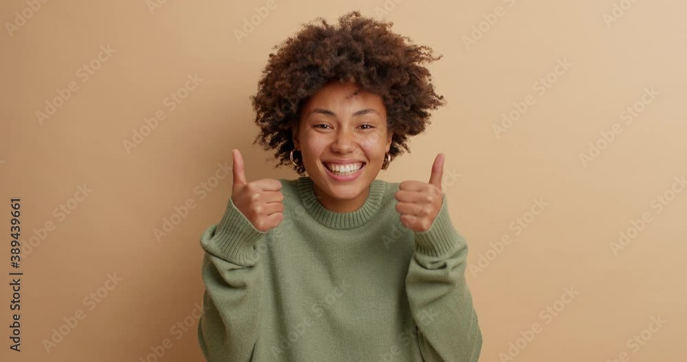 Wall mural happy energized curly woman expresses joy smiles gladfully and raises thumb up shows excellent sign 