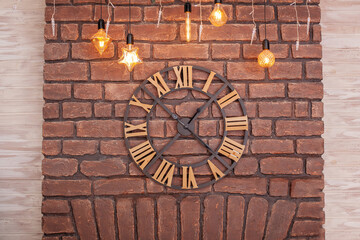 Large classic Clock with roman numerals on a red brick wall. Vintage Clock on wall with glowing garland. Interior concept. Metal wall clock on the background of grunge wall in the Loft apartment.
