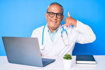 Senior handsome man with gray hair wearing doctor uniform working using computer laptop smiling doing phone gesture with hand and fingers like talking on the telephone. communicating concepts.