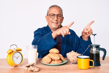 Senior handsome man with gray hair sitting on the table eating breakfast in the morning smiling and looking at the camera pointing with two hands and fingers to the side.