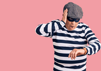 Senior handsome man wearing burglar mask and t-shirt looking at the watch time worried, afraid of getting late