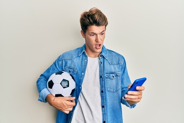 Handsome caucasian man holding football ball looking at smartphone in shock face, looking skeptical and sarcastic, surprised with open mouth