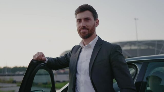 Outdoor portrait of caucasian optimistic young bearded businessman leaning on car door smiling for camera. Successful adult. Business people and transportation.