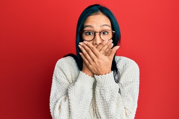 Beautiful hispanic woman wearing casual sweater and glasses shocked covering mouth with hands for...