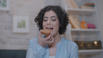 A cheerful young woman is sitting at home on an armchair and eating peanut butter bread.