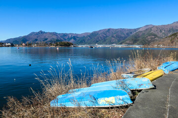 A panoramic view on Kawaguchiko Lake. There are few boats on the shore, turned upside down. The lake shore is overgrown with golden grass. Idyllic landscape. Mountains surrounding the lake. Serenity