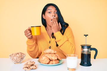 Beautiful latin young woman with long hair sitting on the table having breakfast covering mouth...