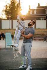 A father helps his son climb a rope on a sports field in masks during sunset. Healthy parenting and healthy lifestyle