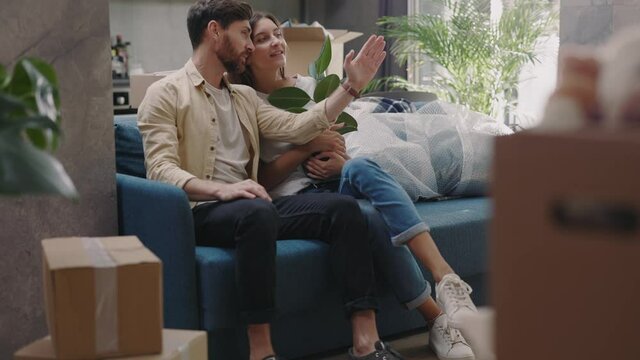 Cute couple move into new apartment. Man and woman sit on sofa planning unpacking stuff smiling talking. Relationships boxes cardboard. At sunlight in modern room. Slow motion
