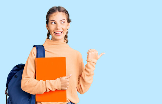 Beautiful caucasian woman with blonde hair wearing student backpack and holding book pointing thumb up to the side smiling happy with open mouth