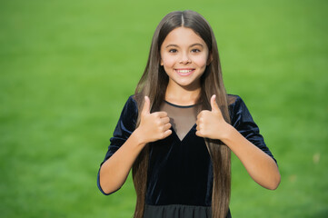 Happy small girl child with long hair and beauty look wear fashion dress showing thumbs ups hands...