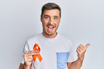 Handsome caucasian man holding awareness orange ribbon pointing thumb up to the side smiling happy...