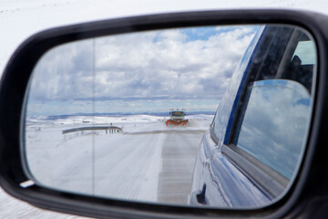 Snow plow cleaning a snow road seen from the rear view mirror of a car.