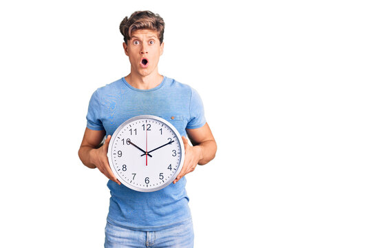 Young handsome man holding big clock scared and amazed with open mouth for surprise, disbelief face