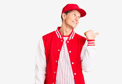 Young handsome man wearing baseball uniform smiling with happy face looking and pointing to the side with thumb up.