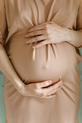 A pregnant woman in a beige dress holds her belly. Vertical close-up.