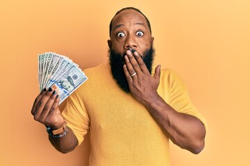 Young african american man holding dollars covering mouth with hand, shocked and afraid for...