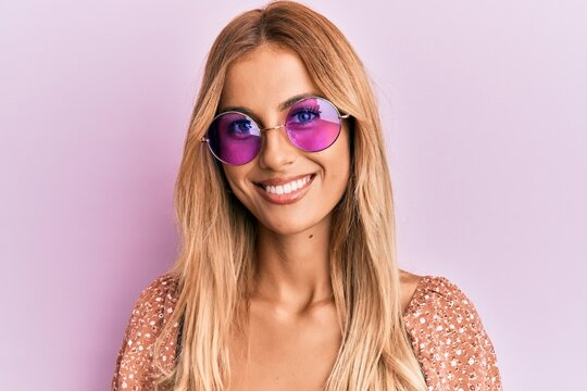 Beautiful blonde young woman wearing fashion pink sunglasses looking positive and happy standing and smiling with a confident smile showing teeth