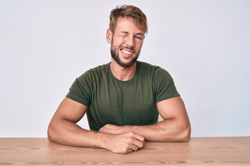 Young caucasian man wearing casual clothes sitting on the table winking looking at the camera with sexy expression, cheerful and happy face.