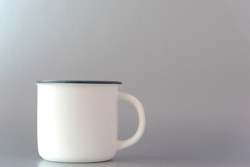 Stylish white metal mug with handle and mockup for template on side stands on light grey background extreme close view. Contemporary dishware for beverages.