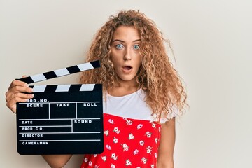 Beautiful caucasian teenager girl holding video film clapboard scared and amazed with open mouth...