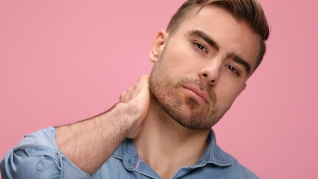 young man in blue shirt sensually holding hands behind neck and head and posing on pink background in studio