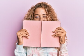 Fototapeta premium Beautiful caucasian teenager girl reading a book covering face winking looking at the camera with sexy expression, cheerful and happy face.