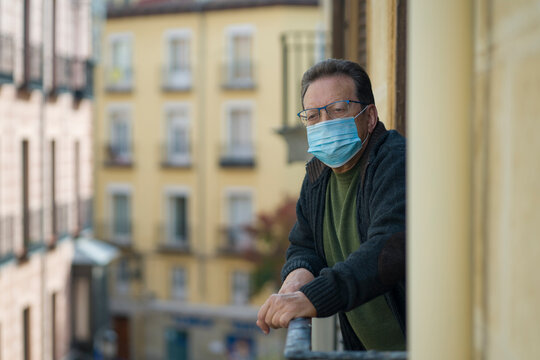 mature man in balcony home lockdown during covid19 outbreak - senior male on his 70s in face mask worried and tired looking to the street thoughtful and depressed in quarantine